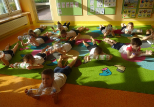 Dzieci leżą na brzuchu z wyciągniętymi rękami przed sobą w których trzymają plastikową butelkę.
